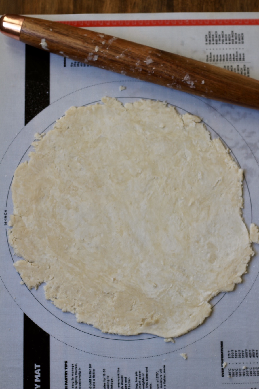 A pastry mat with a 12 inch round rolled out pie crust dough and a wooden rolling pin on top.