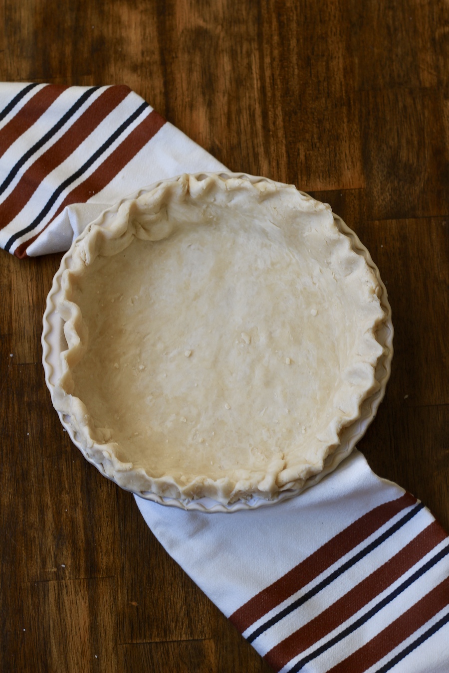 A pie pan with uncooked pastry crimped at the edges on a wooden counter with a white and brown dish towel.