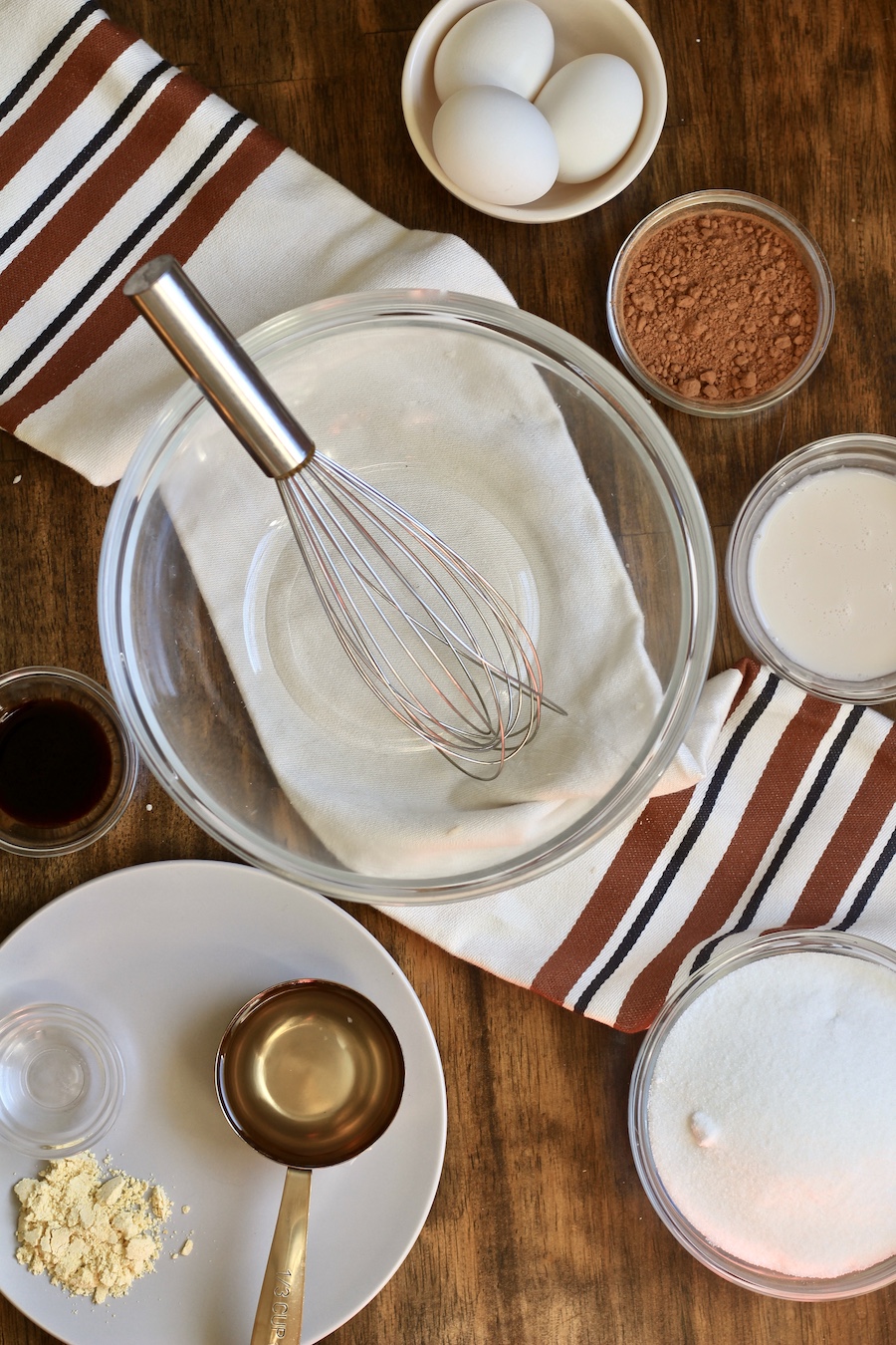 A glass mixing bowl with a metal whisk in the middle with chocolate chess pie ingredients around it on a wooden counter with a white and brown dish towel.