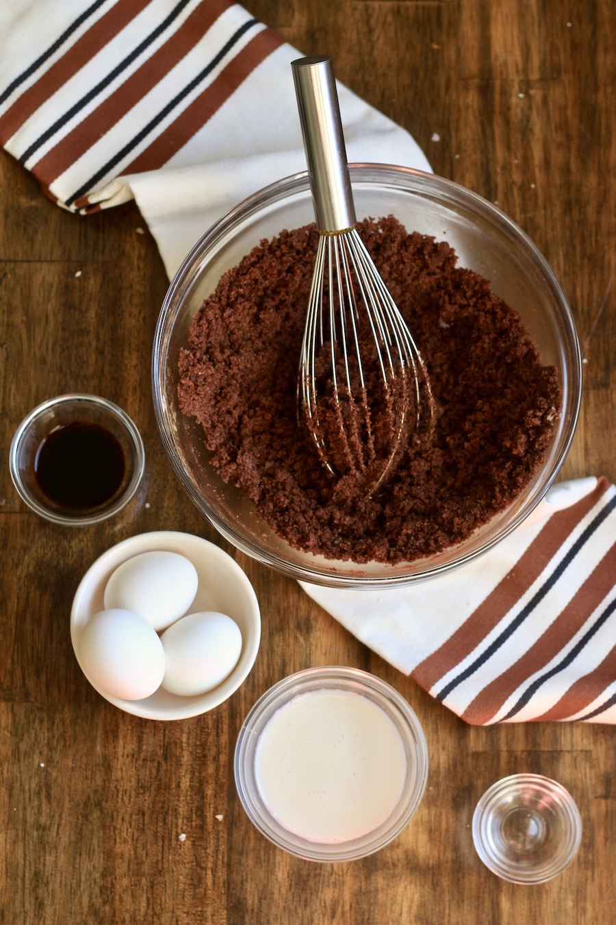 A glass bowl with some chocolate chess ingredients mixed in and the rest of the ingredients in front on a wooden table with a white and brown dish towel.