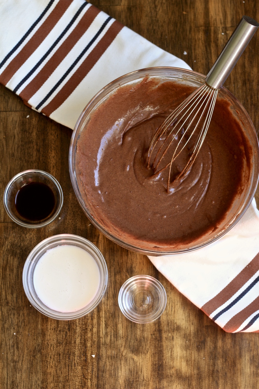 A glass mixing bowl with chocolate chess pie batter and a silver whisk with non-dairy milk, vinegar, and vanilla in small bowls in front on a wooden counter with a white and brown dish towel.