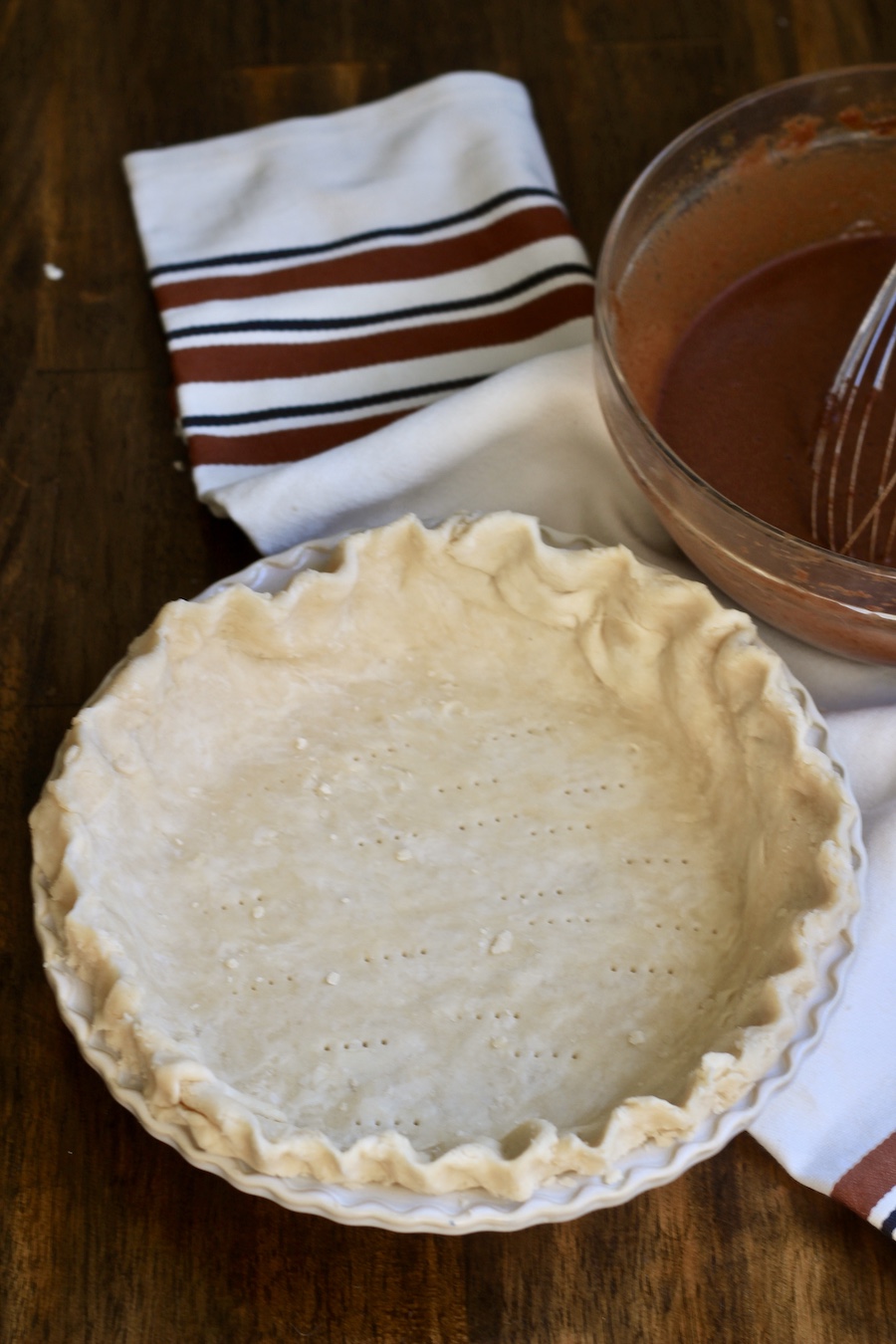 An uncooked pie crust dough in a pie pan with a glass mixing bowl in the back with chocolate chess pie filling and a whisk on a wooden counter with a white and brown dish towel.