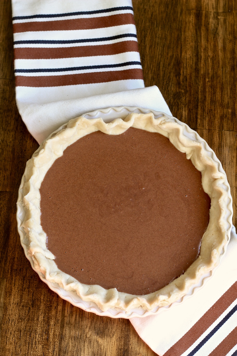 Chocolate chess pie in a pie pan before baking on a white and brown dish towel on a wooden counter.