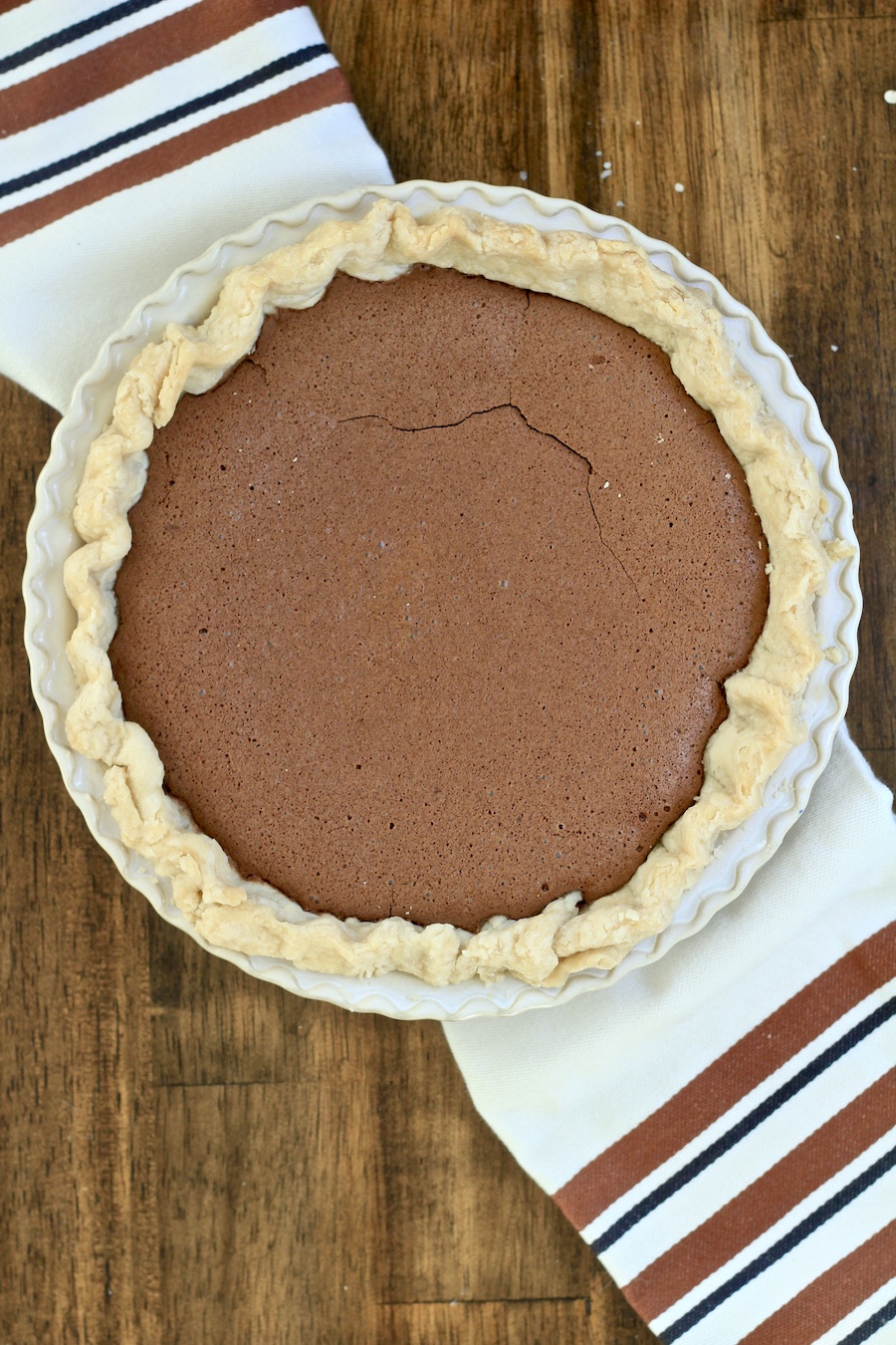 A baked chocolate chess pie on a white and brown dish towel on a wooden counter.