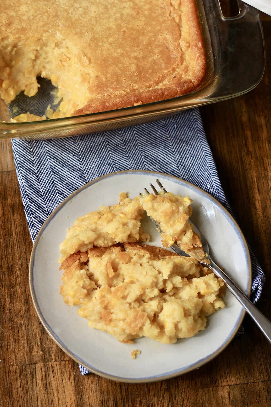 A scoop of corn casserole on a small plate with a fork on a blue dish towel with a baking dish of corn casserole in the back.