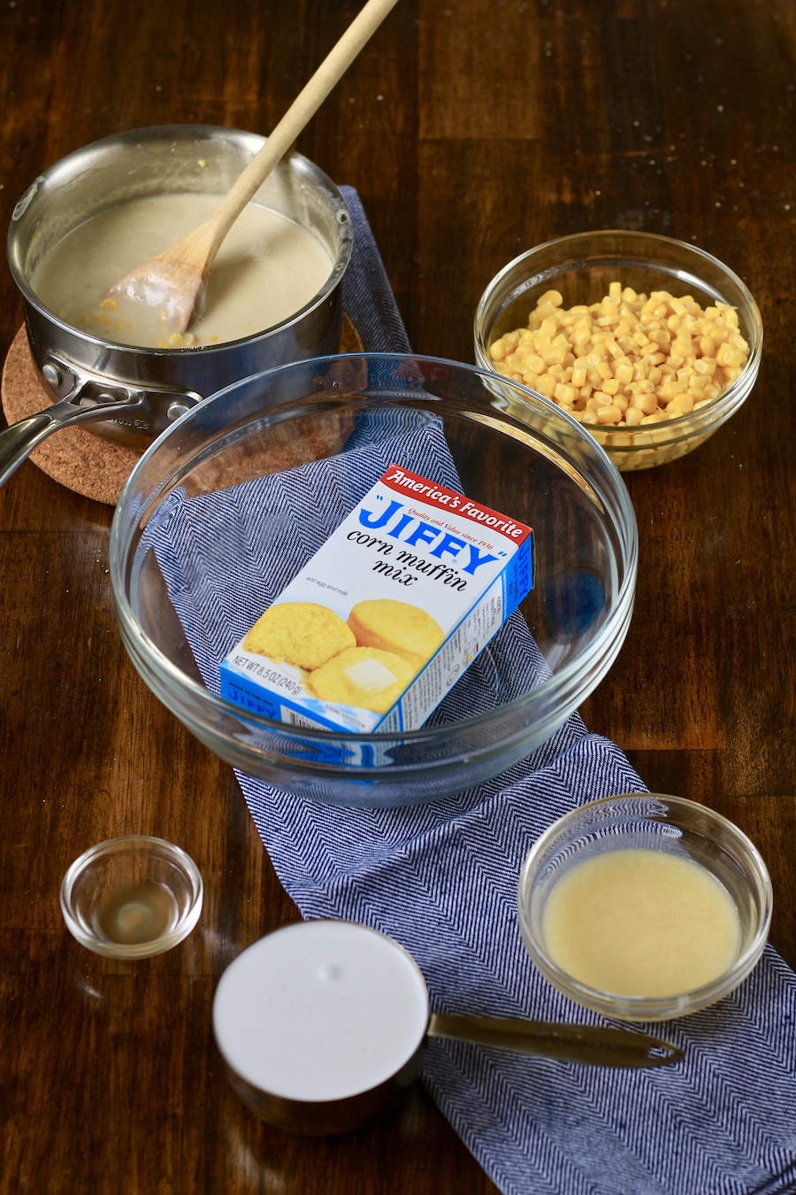Ingredients for corn casserole on a wooden table in the top left a vegan creamed corn next to a bowl of drained corn, with a glass bowl with a box of jiffy corn muffin mix in the middle, and a bowl of melted vegan butter in the bottom right, a measuring cup of coconut milk in the bottom center, and spiced on the bottom left.