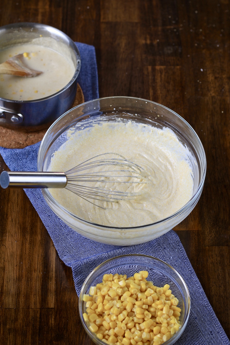 A mixing bowl in the middle with corn bread mixed with a whisk, a bowl of corn at the bottom and a pot of vegan creamed corn at the top.