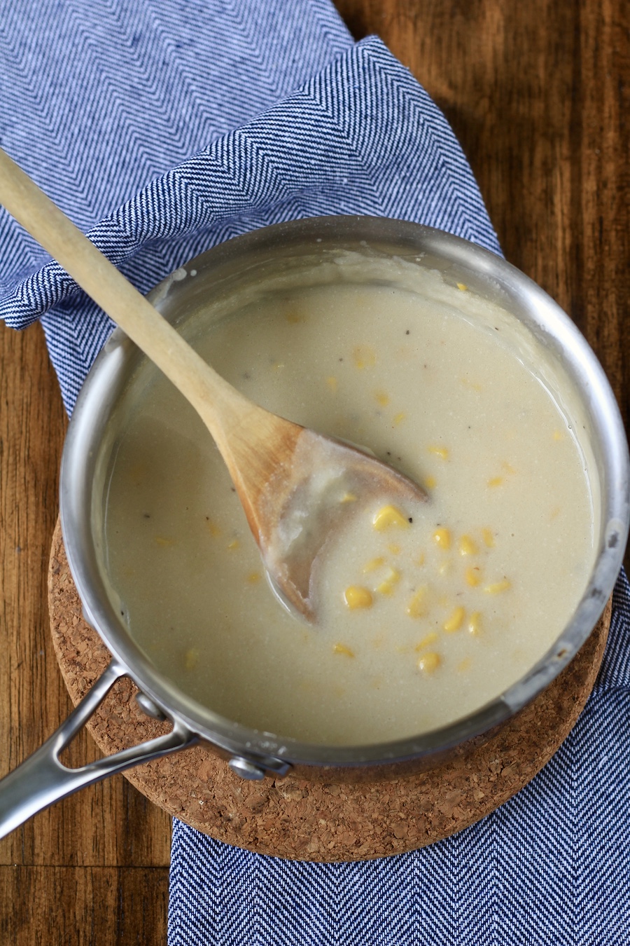 A sliver pot with vegan creamed corn with a wooden spoon on a cork trivet on a blue towel on a wooden counter.