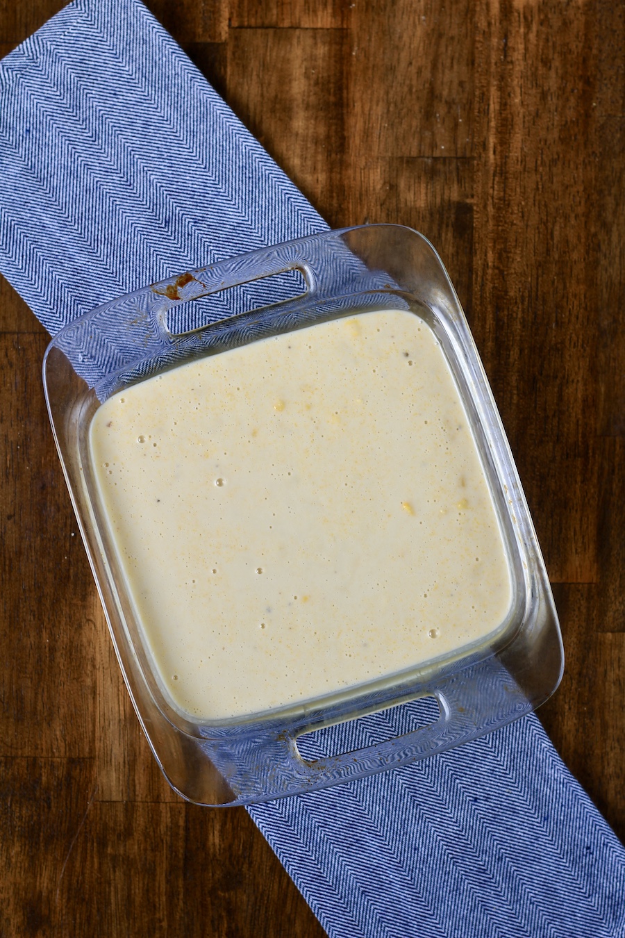 Unbaked corn casserole in a square baking dish on a blue towel on a wooden counter.