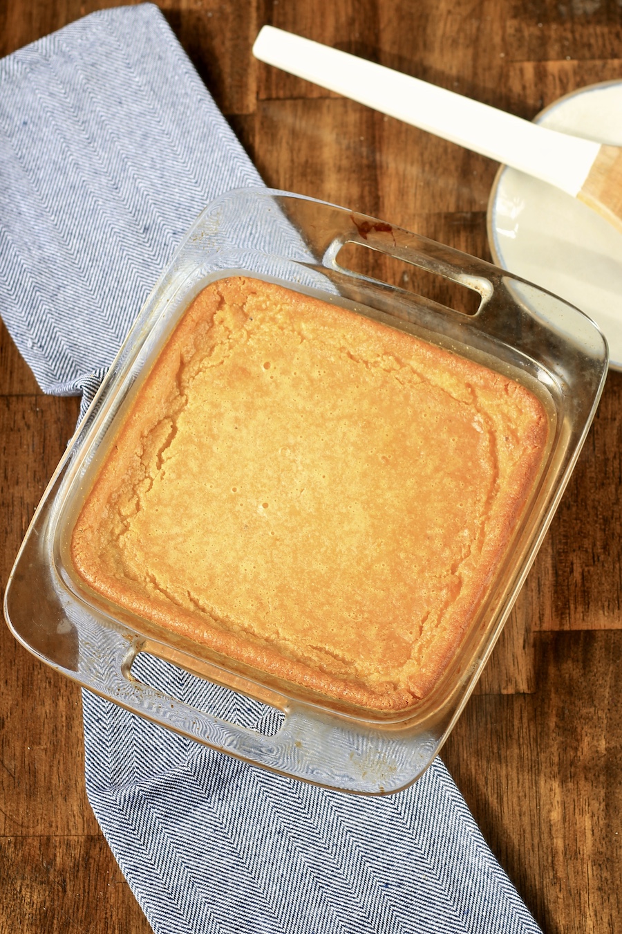 Baked corn casserole on a blue towel with a small plate and spoon in the background on a wooden counter.