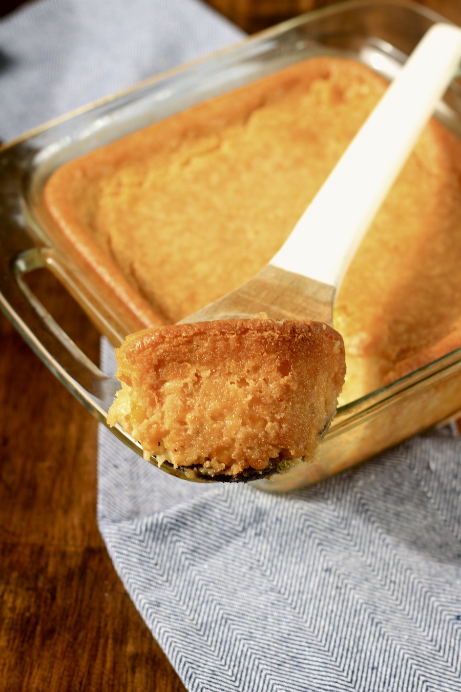 A scoop of corn casserole on a wooden spoon on top of the baking dish of corn casserole on a blue and white towel in a wooden counter.