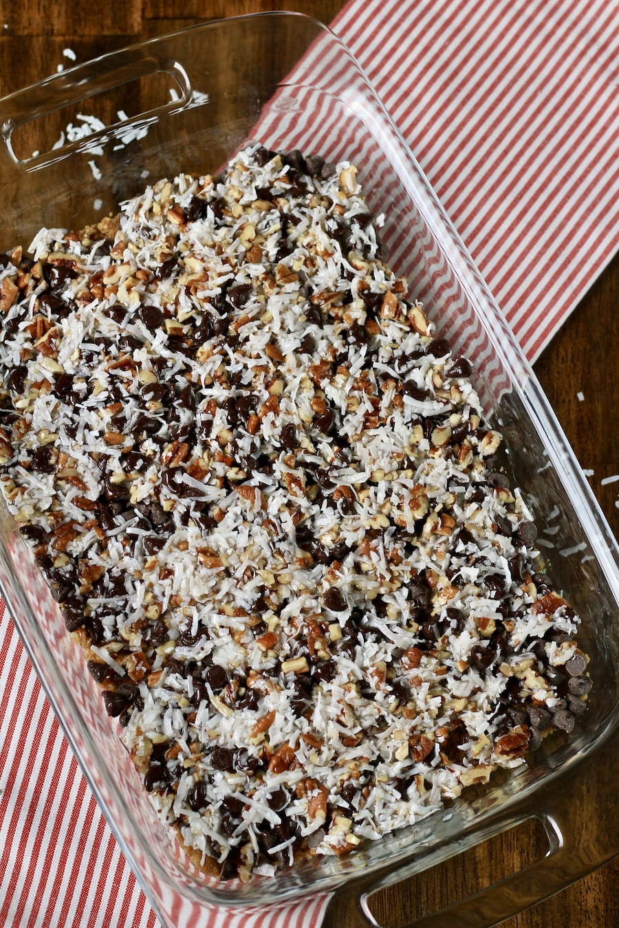 A glass baking dish with unbaked magic cookie bars on a red and white striped towel on a wooden table.