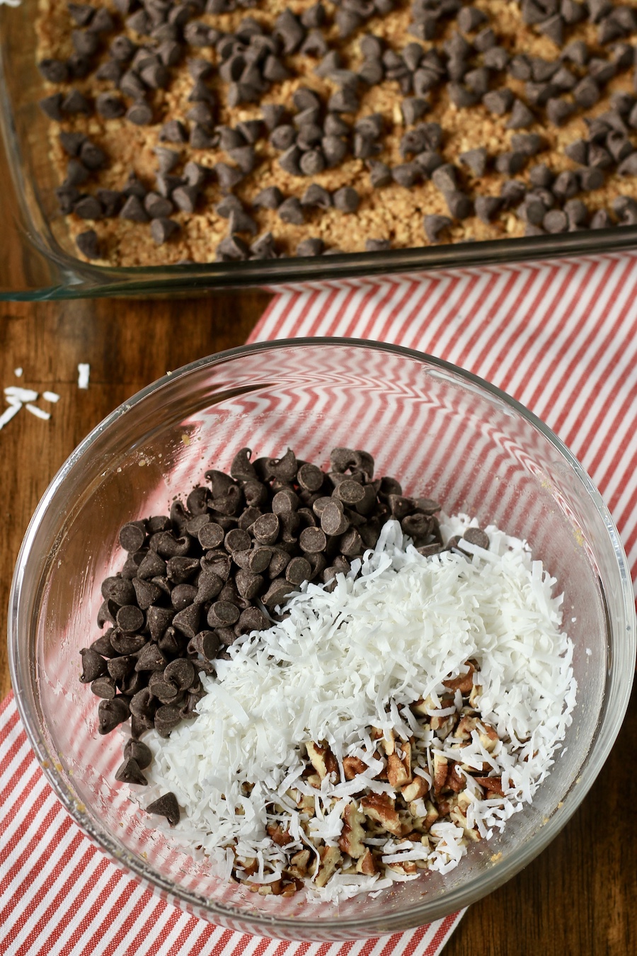 A glass mixing bow with chocolate chips, coconut shreds, and chopped pecans on a red and white striped towel with a glass baking dish with a graham cracker crust topped with chocolate chips.