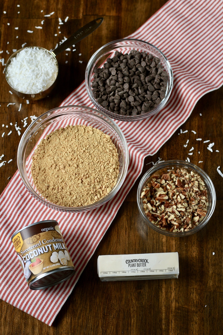 A glass bowl with coconut shreds, a bowl with chocolate chips, a bowl with graham cracker crumbs, a bowl with chopped toasted pecans, a can of sweetened condensed coconut milk, and a stick of vegan butter on a red and white dish towel.