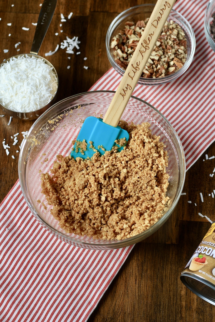 A glass bowl with graham cracker crumbs mixed with melted butter and a teal spatula in the bowl with a small bowl of coconut shreds and chopped pecans in the back.
