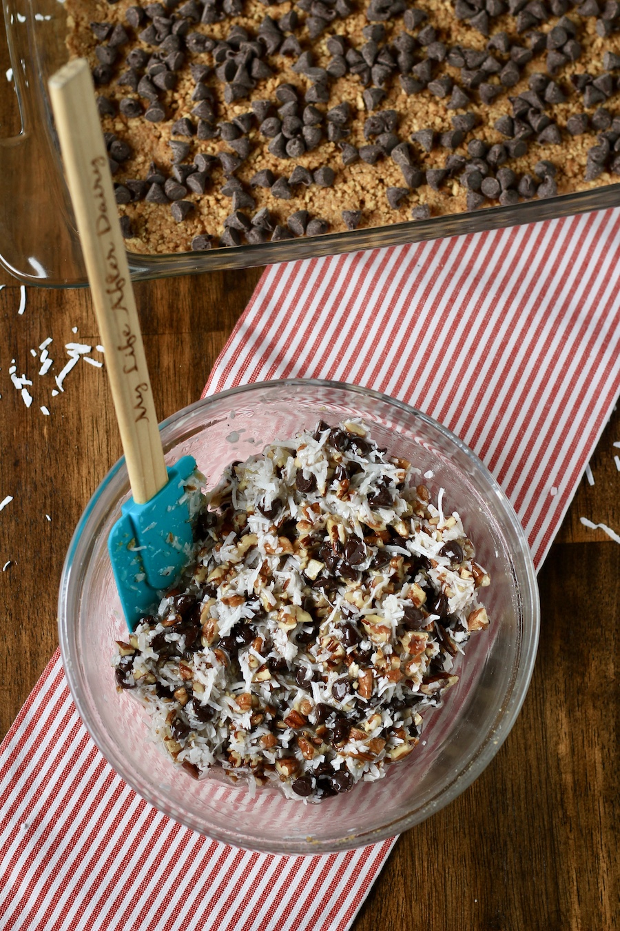 A glass bowl with coconut shreds, toasted pecans, and chocolate chips mixed with sweetened condensed coconut milk on a red and white dish towel in front of a baking dish with a graham cracker crust topped with chocolate chips.