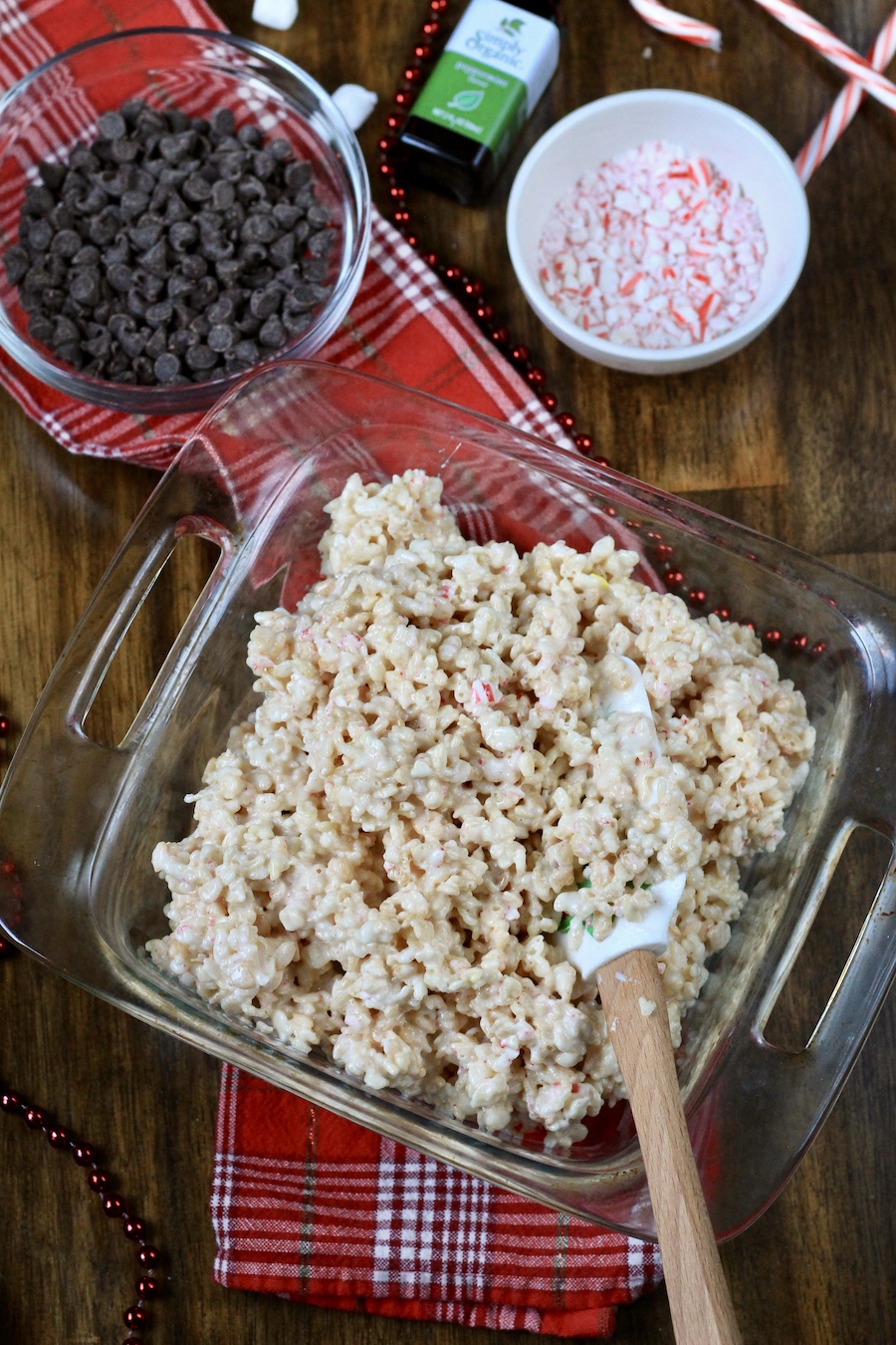 A square glass baking dish with rice krispie treats unpressed in the pan with chocolate, peppermint, and peppermint extract behind the baking dish.