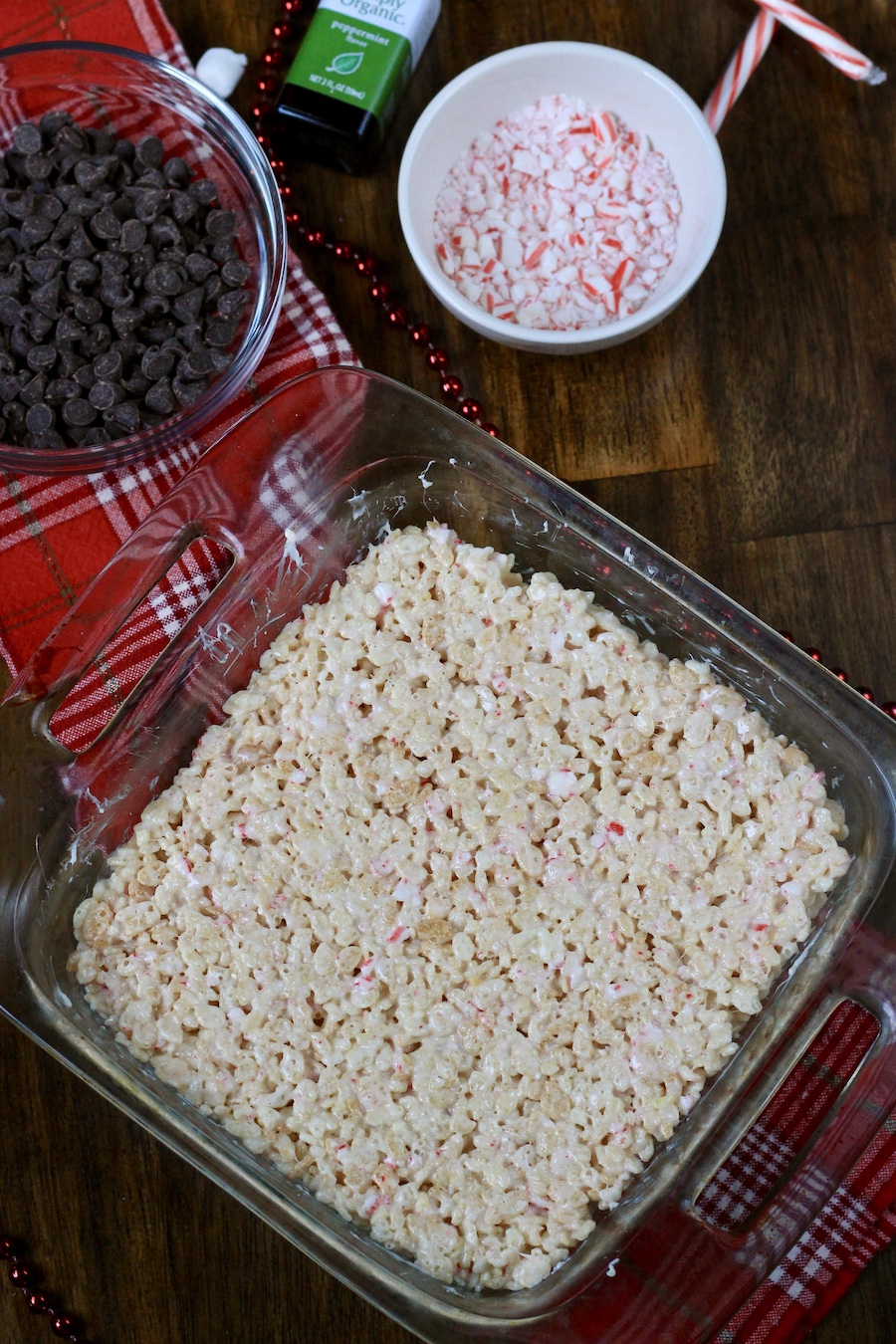 Peppermint rice krispies pressed into a square baking dish with ingredients for peppermint bark topping behind.