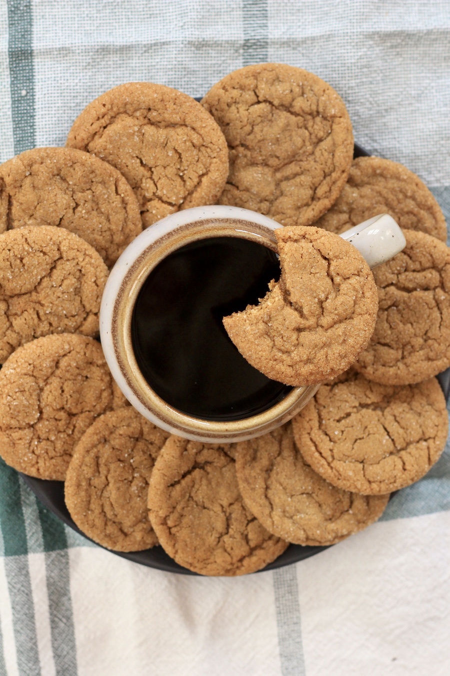 A ring of ginger cookies around a coffee cup with a cookie on top of the mug with a bite taken out on top of a green and white dish towel.