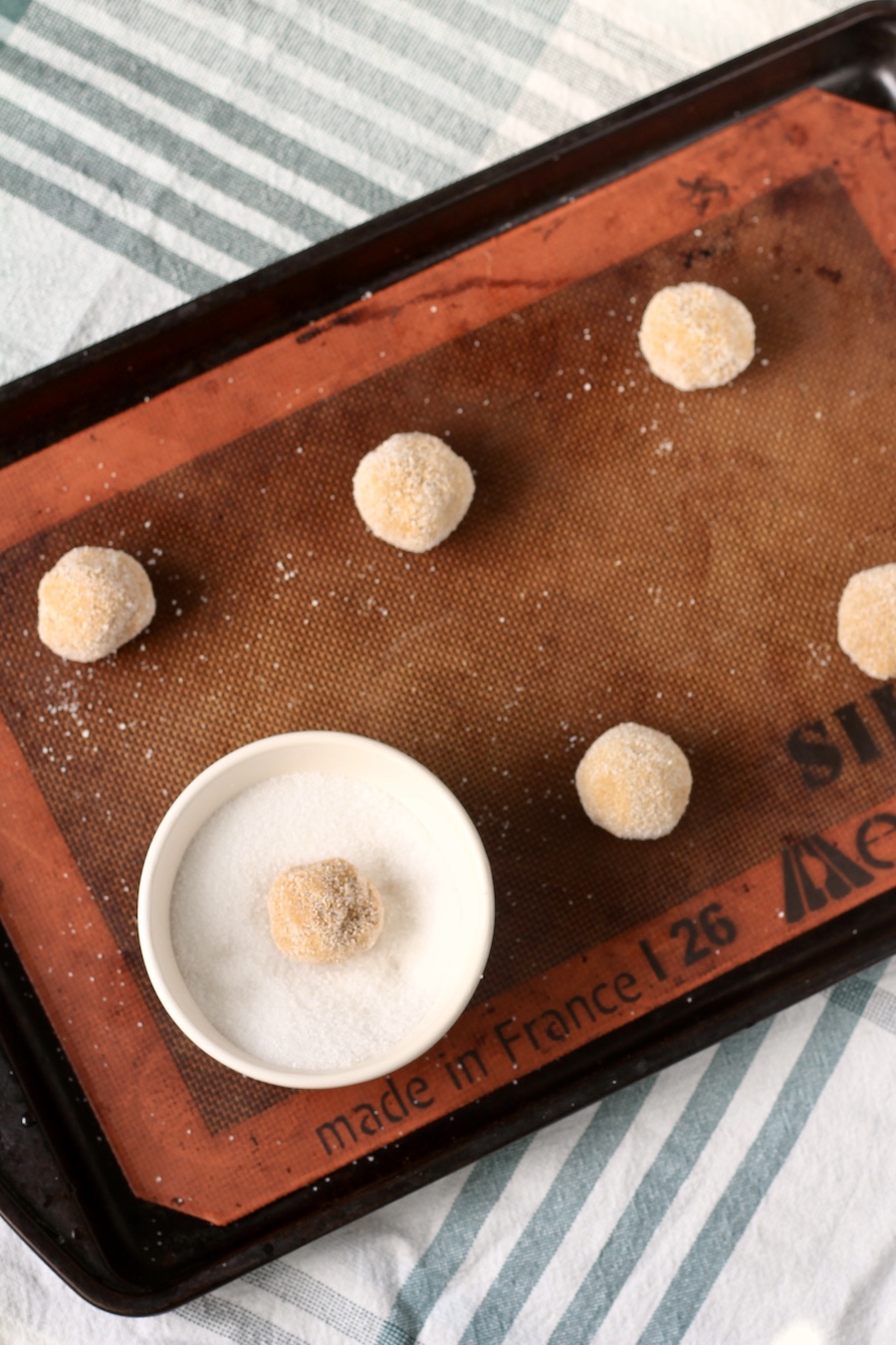 Balls of soft ginger cookie dough rolled in sugar on a baking sheet with a silpat on a white and green towel.
