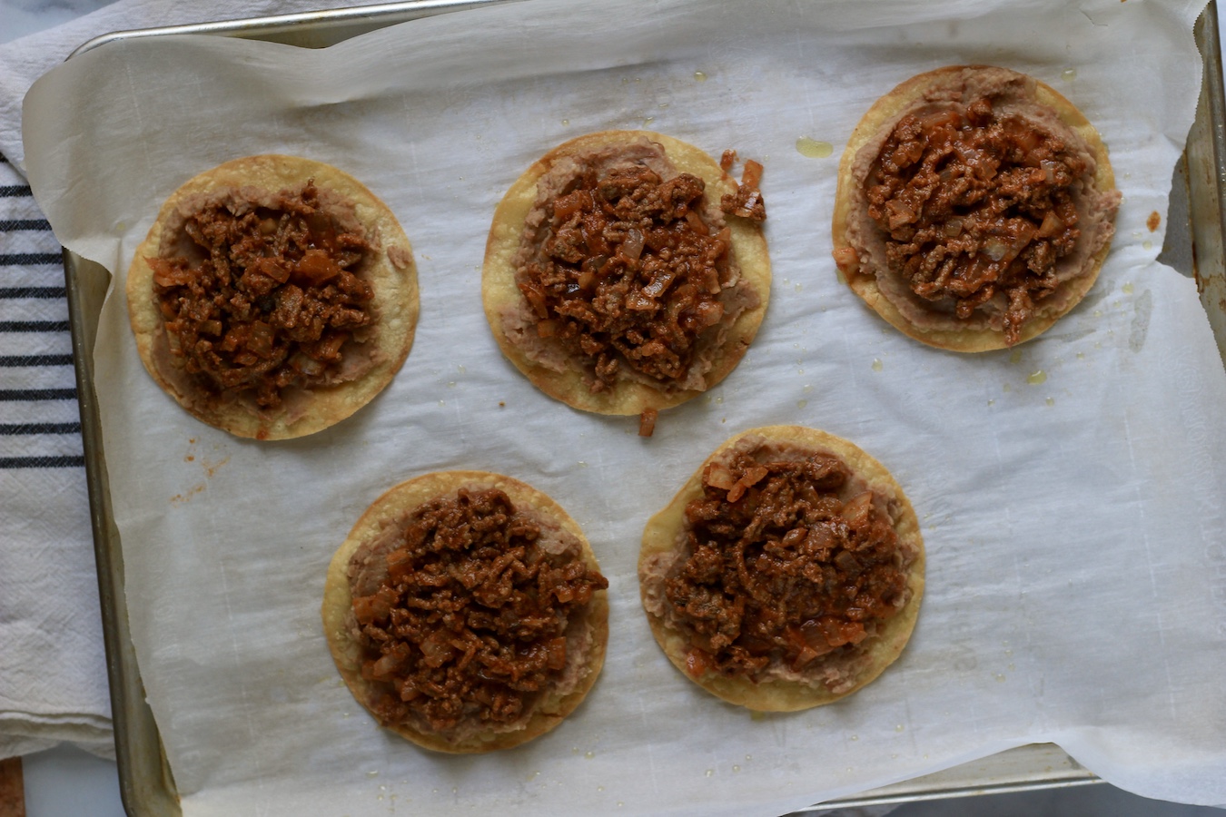 A baking sheet lined with parchment paper with corn tortillas topped with refried beans and beef taco meat.