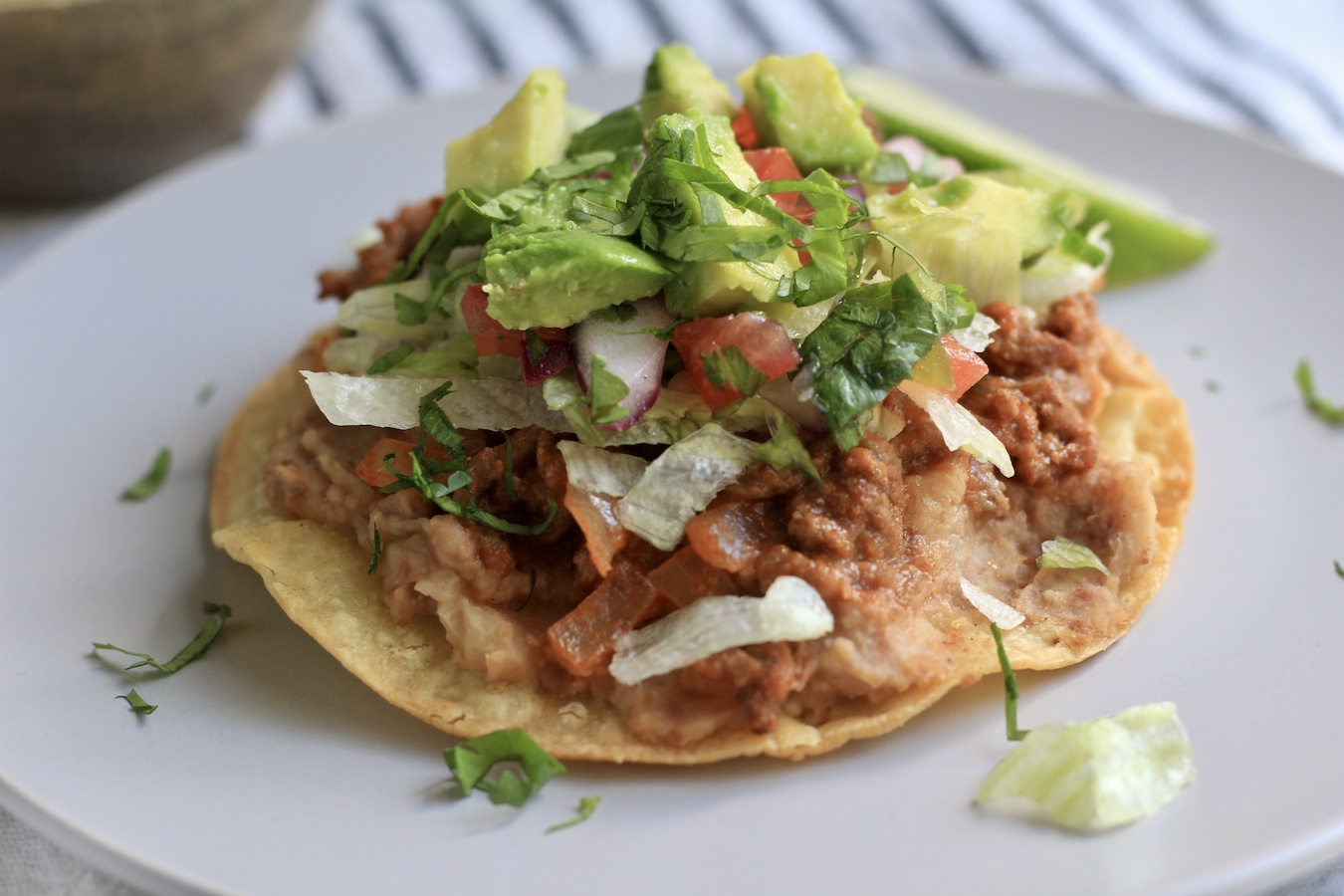 An oven baked beef tostada on a white plate topped with avocado and shredded lettuce.