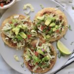 Three oven baked beef tostadas on a white plate with a lime wedge and a small bowl of pico de gallo in the back.