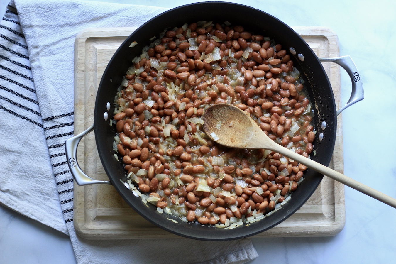 Beans cooking in a non-stick skillet with a wooden spoon on a wooden cutting board.