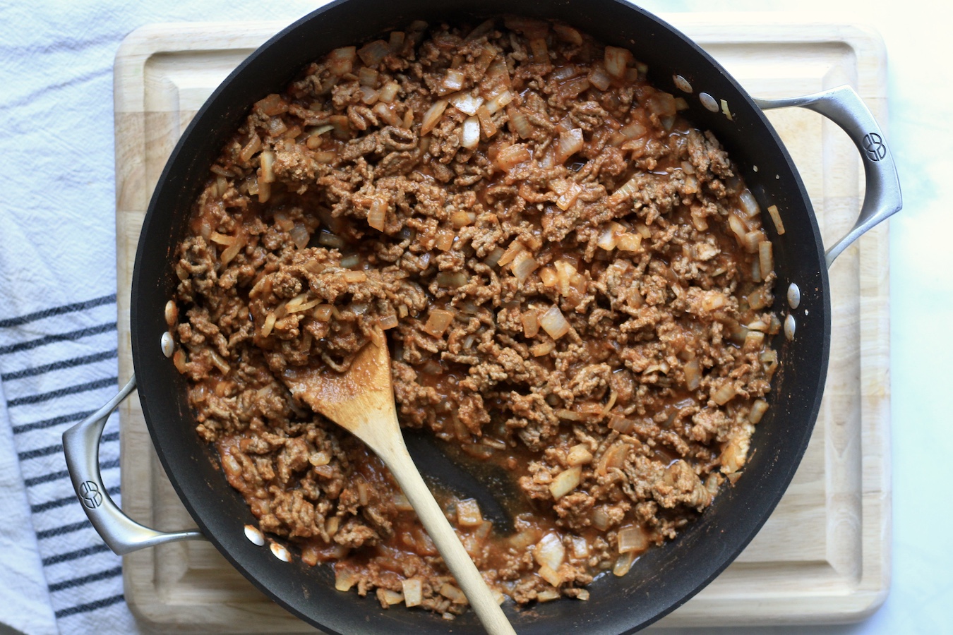 Beef taco meat in a non-stick skillet with a wooden spoon on a wooden cutting board.
