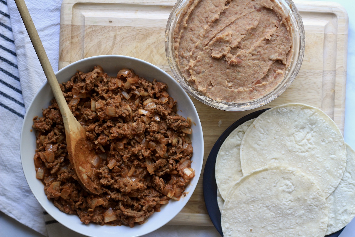 A white bowl on the left with beet taco meat, a small bowl of creamy refried beans at the top, and a plate with corn tortillas on the right.