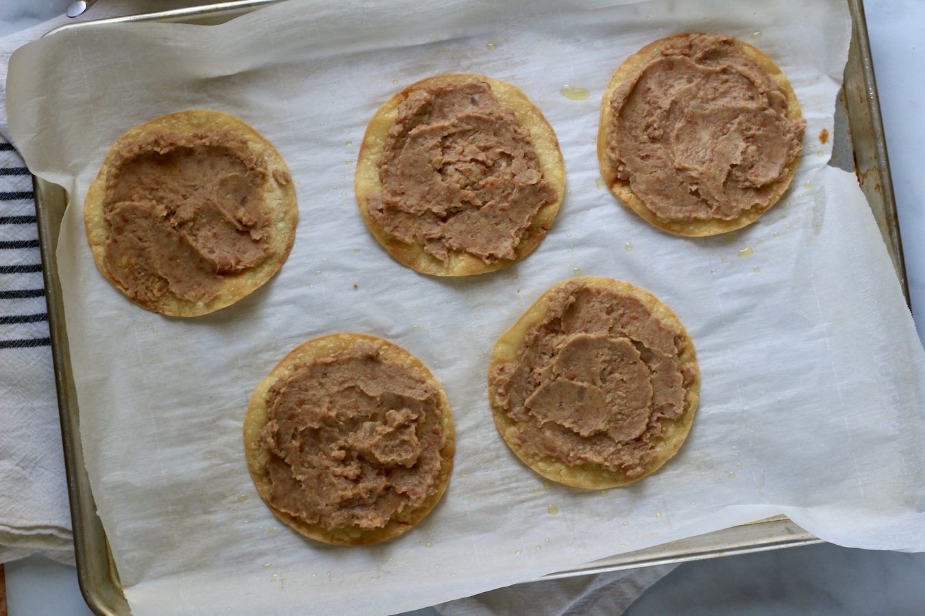 A baking sheet lined with parchment paper with corn tortillas topped with refried beans.