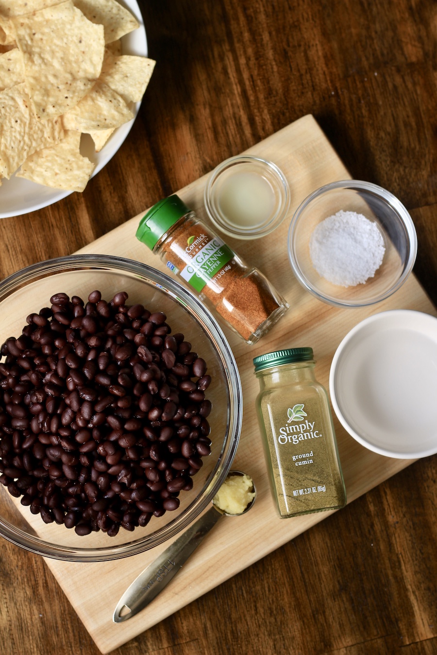 A wooden table with a wood cutting board with beans, spices, and water on the cutting board.