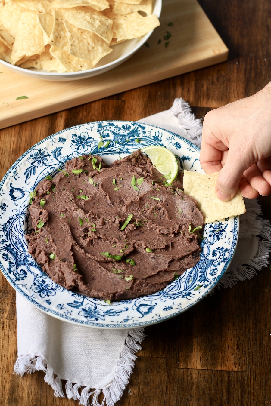 A hand dipping a tortilla chip into black bean dip in a white and blue bowl on a white towel.