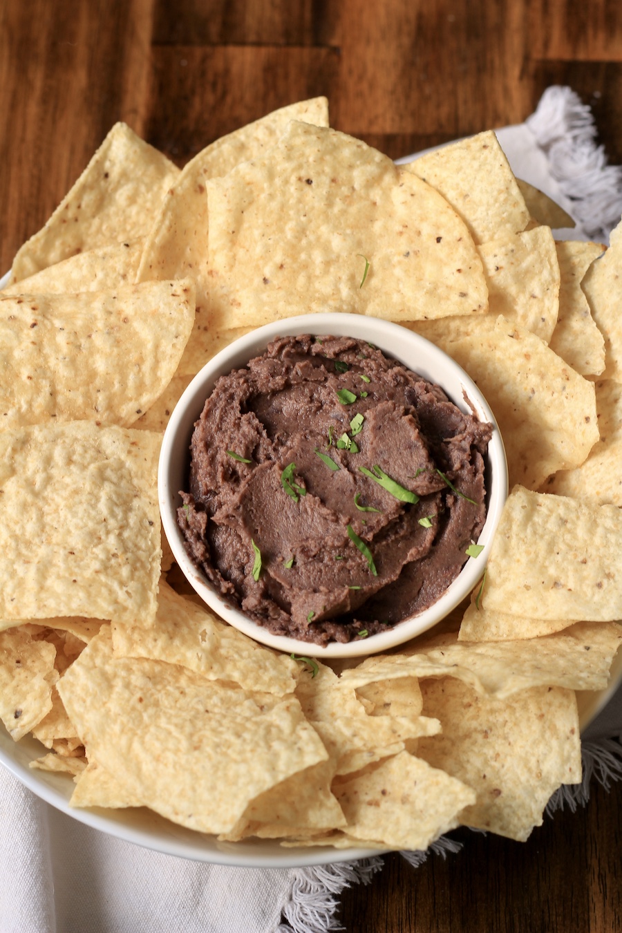 A small bowl of black bean dip in the middle of a big bowl of tortilla chips on a wooden counter.
