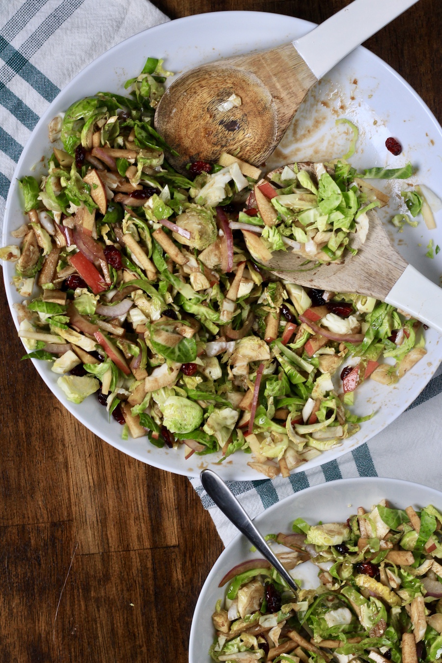 A large white bowl with Brussels Sprouts salad on a white and green dish towel with serving spoons.