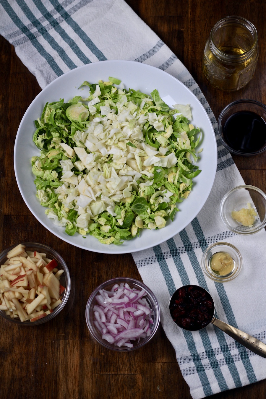 A white bowl with chopped brussels sprouts and cabbage with glass bowls of salad ingredients on a wooden counter with a white and green towel.