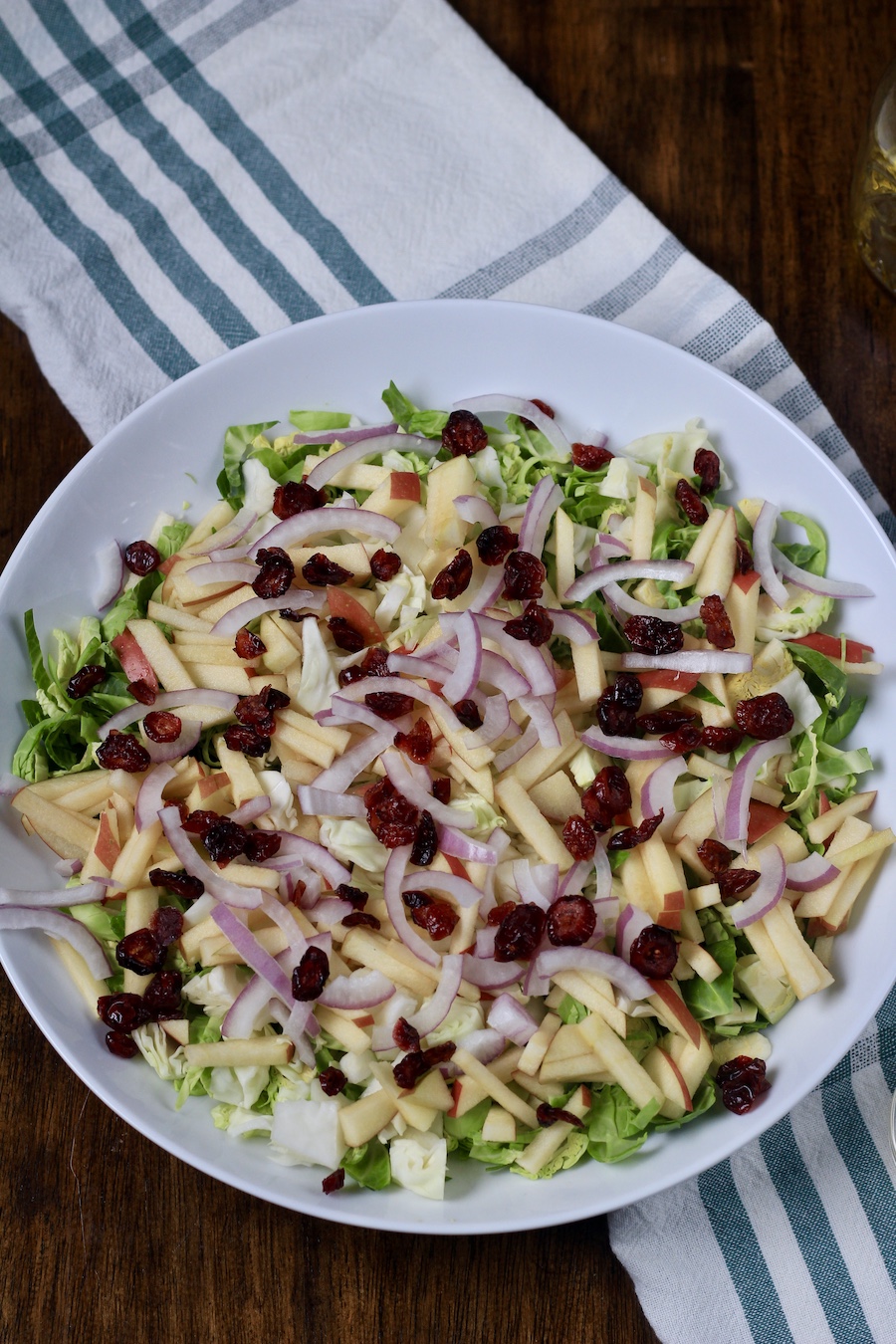 Brusseles sprouts salad topped with craisins on a wooden counter with a white and green dish towel.