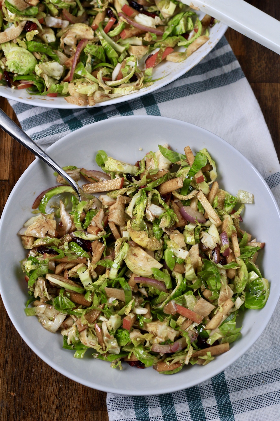 A serving bowl with brussels sprouts salad with a fork in the top on a white and green dish towel.