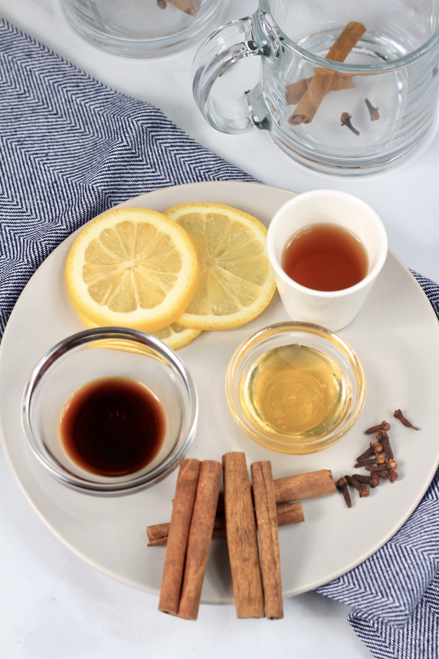 A white plate with the ingredients for hot toddies measured in bowls with mugs in the back and a blue dish towel.