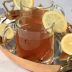A glass mug of a hot toddy with slices of lemon in a copper platter with lemon.