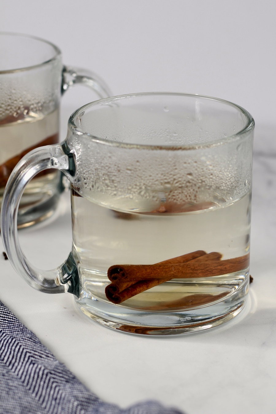 A clear mug with water and cinnamon sticks on a white counter with a blue dish towel in the front.