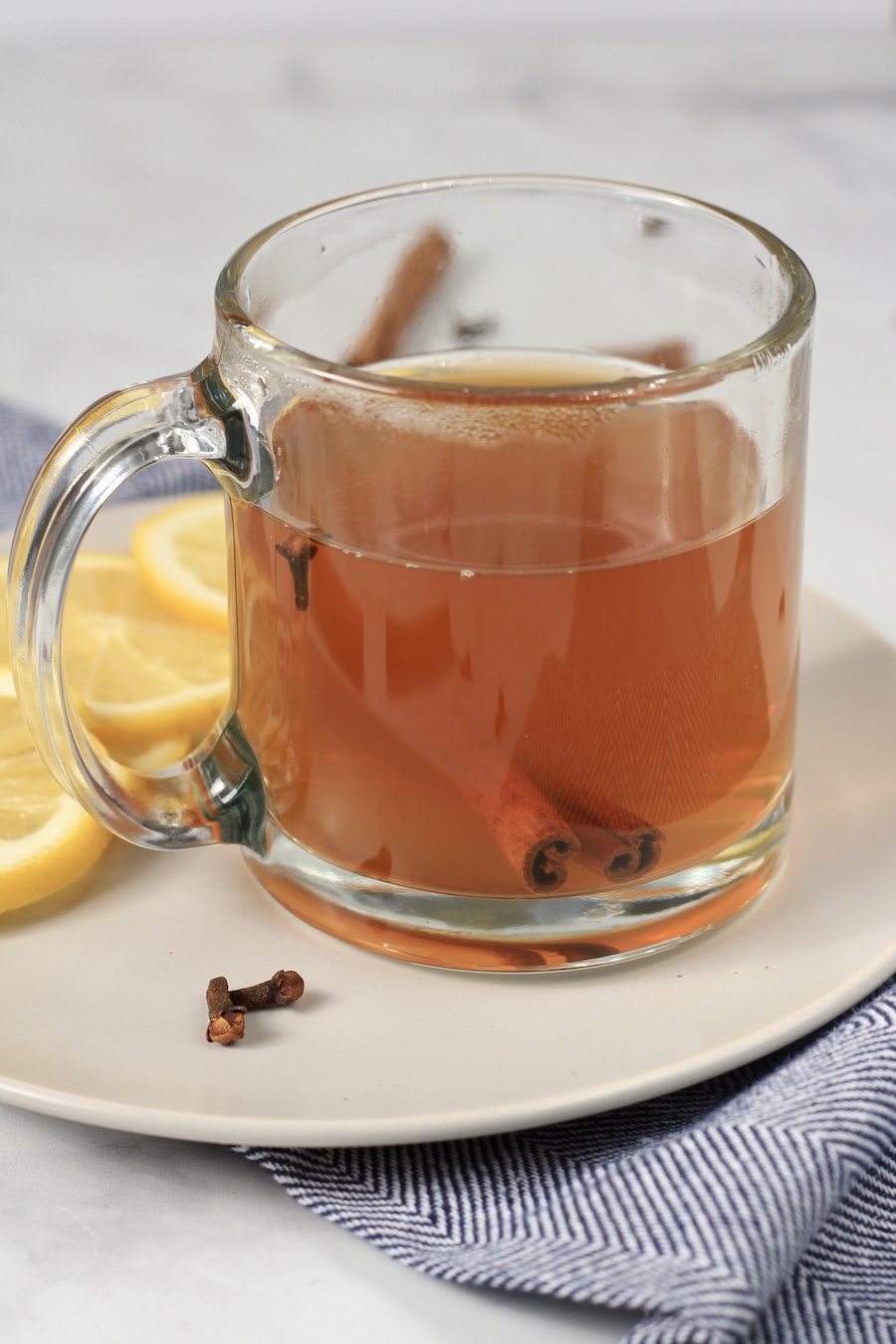 A white plate with a clear glass mug filled with a hot toddy and slices of lemon in the back.