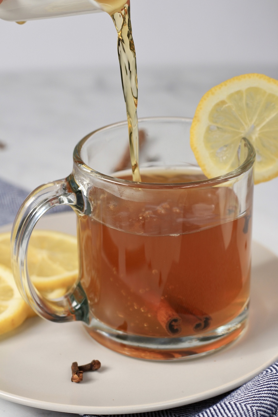 Whiskey being poured into a hot toddy with a lemon slice on the mug and slices in the back on a white plate.