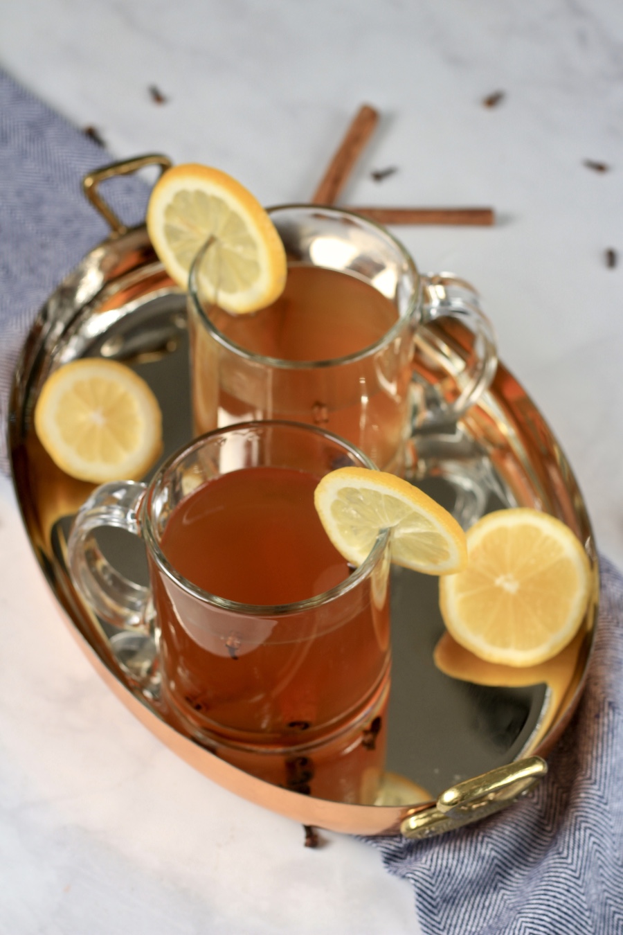 A copper platter with two mugs of hot toddy with slices of lemon on the edges and some cinnamon sticks in the back.