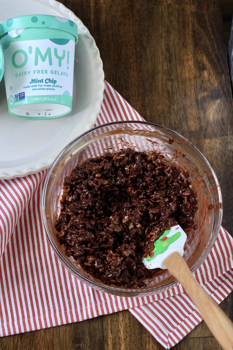 A clear mixing bowl with the chocolate coated rice krispies and a rubber spatula on a red and white striped towel.