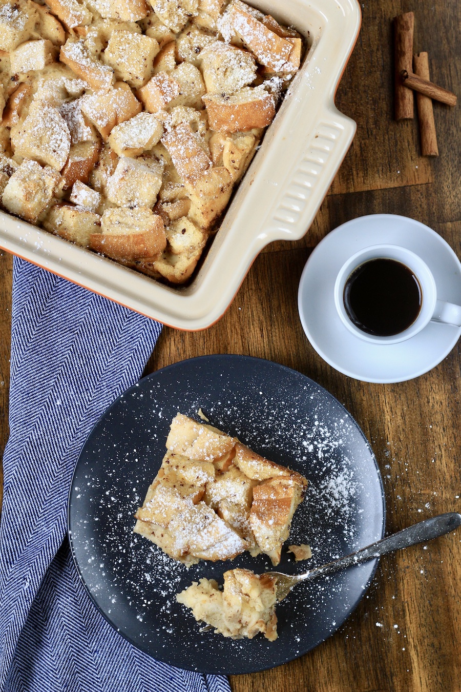 A baking dish with vanilla bread pudding at the top, a cup of coffee in the middle right, and a blue plate with a serving of bread pudding with a fork in the front.
