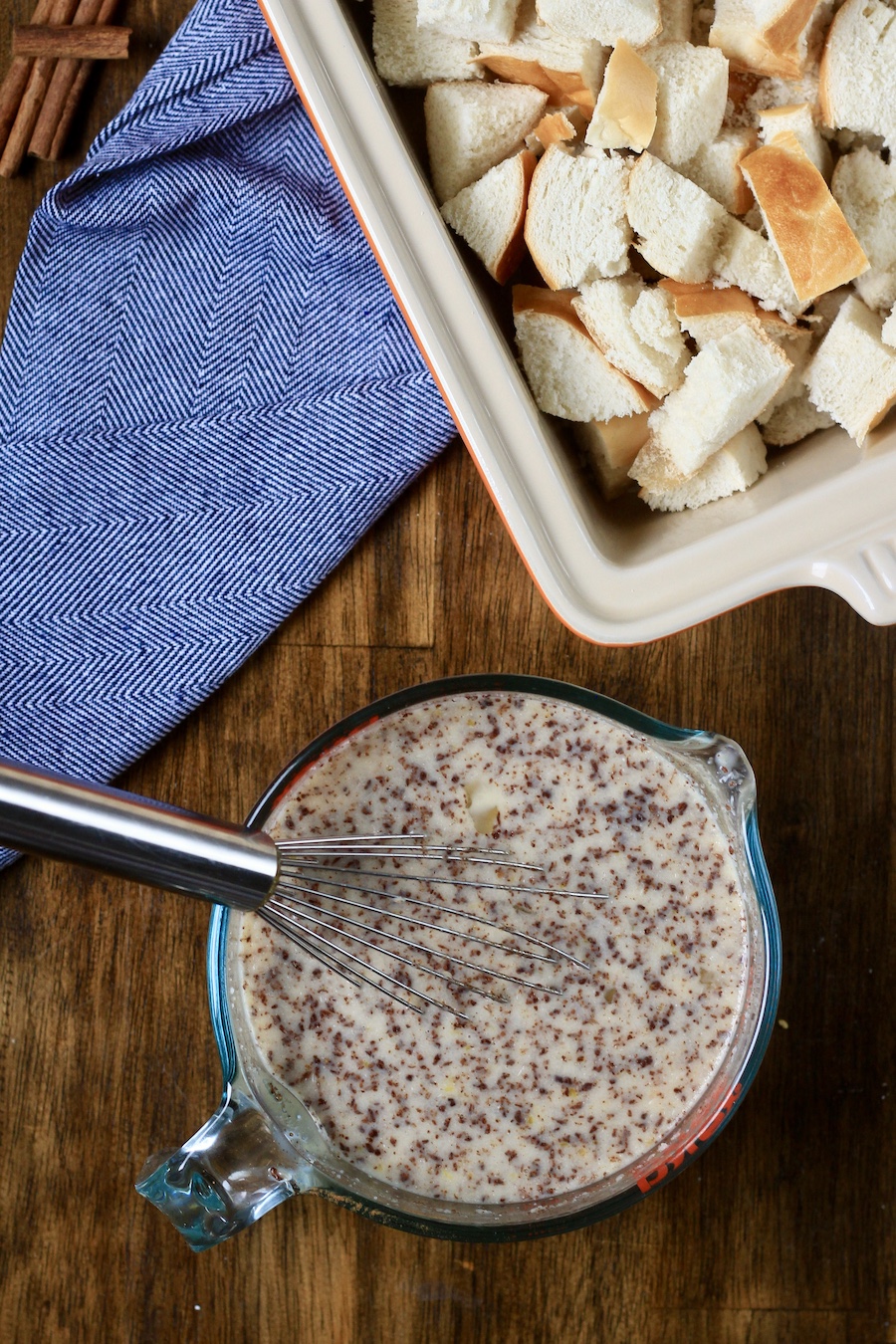 A liquid measuring cup with the liquid ingredients and a metal whist in front of the cubed bread in a baking dish.