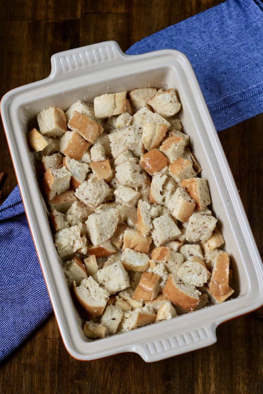 The vanilla bread pudding in a baking dish after being coated with the liquid ingredients on a wooden table with a blue towel.