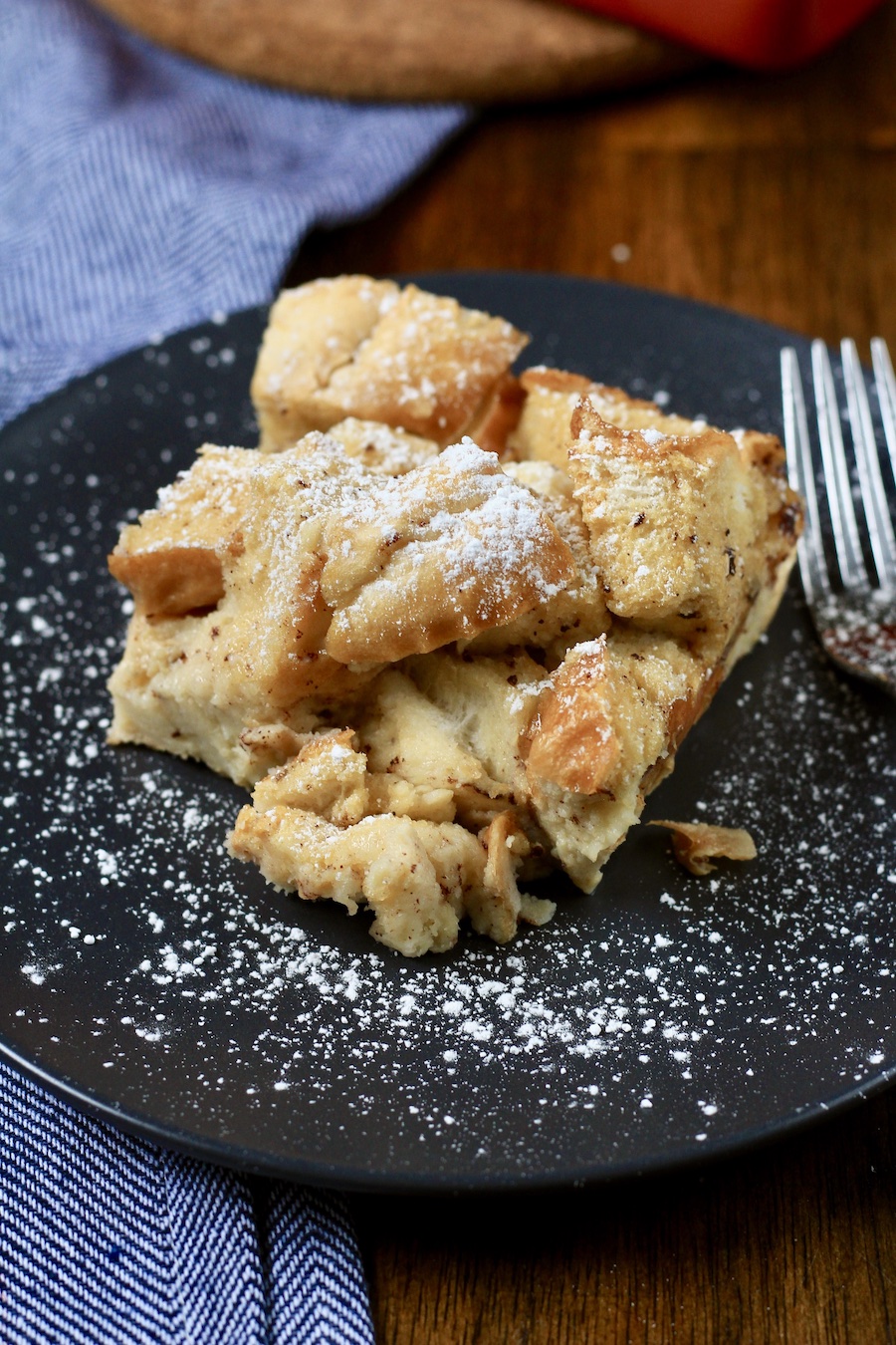 A blue plate with a cut of vanilla bread pudding dusted with powdered sugar with a metal fork to the right.