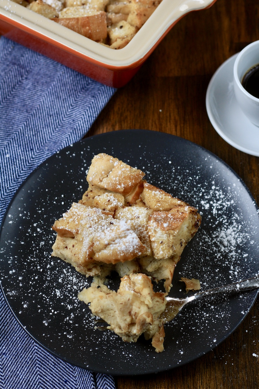 A blue plate with a serving of vanilla bread pudding with a fork in front and a cup of coffee in the back.